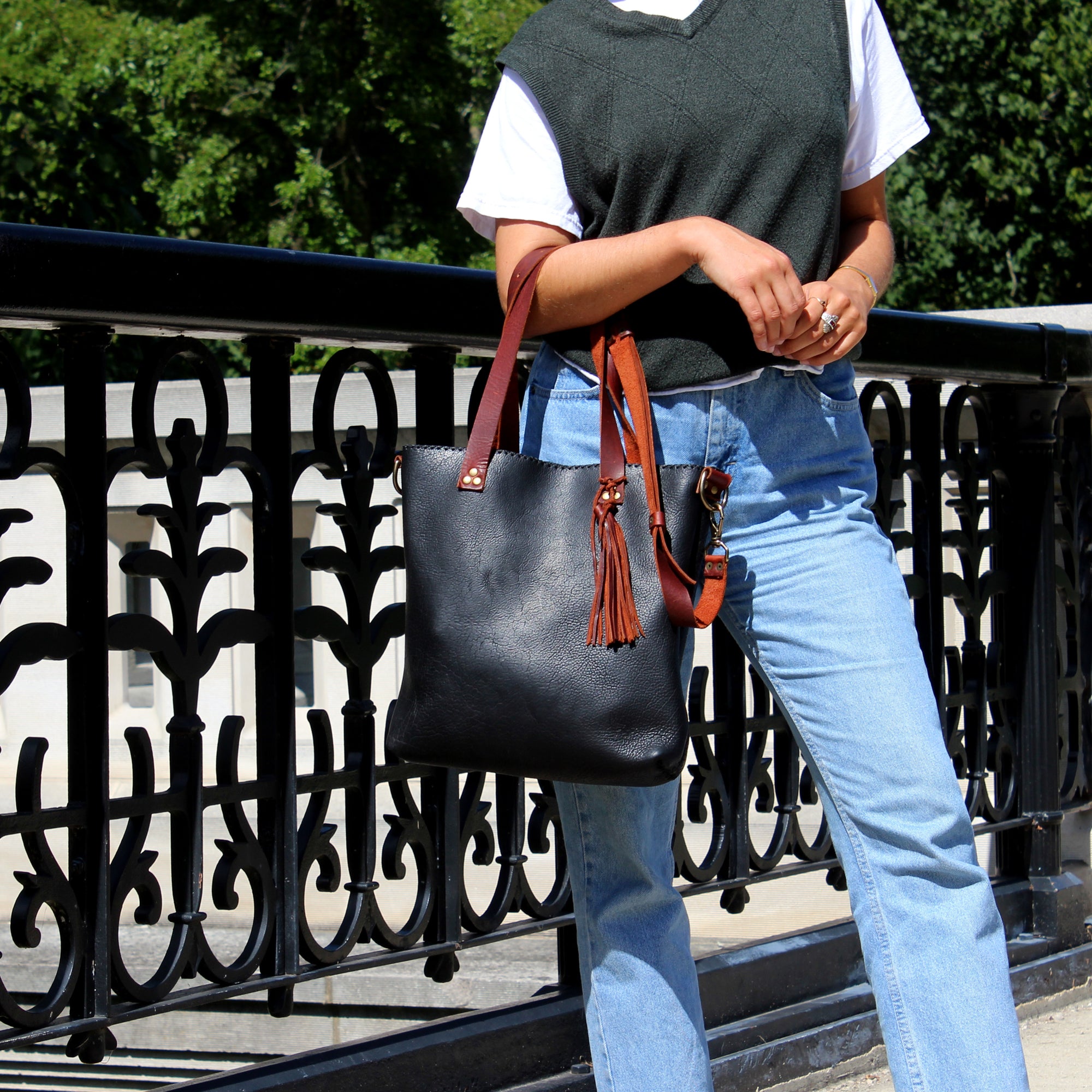 The body of a woman in blue jeans and a green sweater vest standing on the side of a bridge. She models a black and brown leather bag.