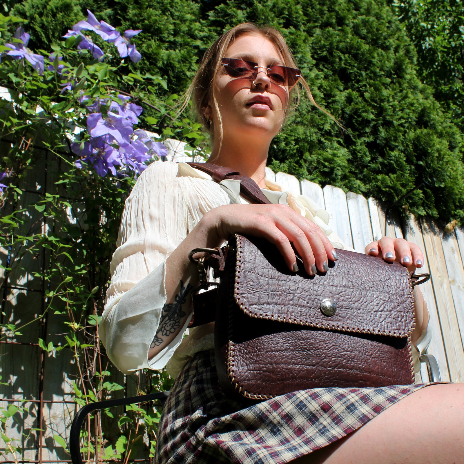 A woman with blonde hair and sunglasses gazes down at the camera while holding a brown leather lag on her lap.