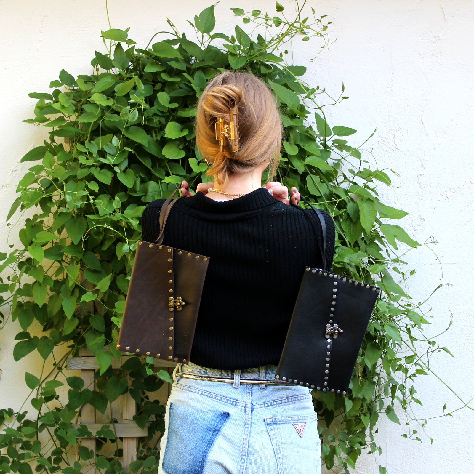 A woman with blonde hair holds two leather clutches by the strap over her back and shoulders.