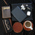 An assortment of accessories laid on on a table. From the top left: a book of poems, glasses case, leather coasters with a coffee cup on it, a tablet, black tablet case, car keys, and headphones.
